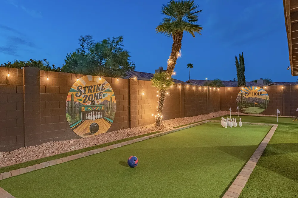 A bowling lane laid on turf beside the garden wall at dusk, pins set and string lights above.