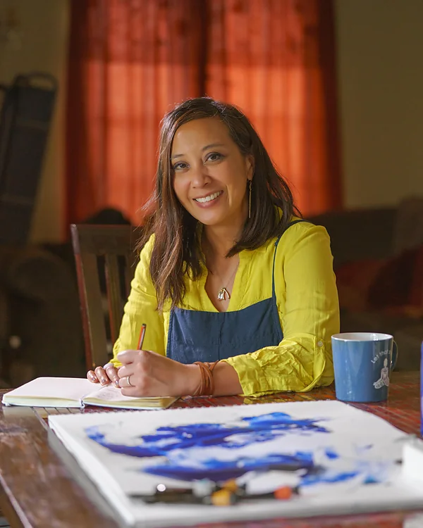 Clarissa Castillo-Ramsey at a table, pencil in hand, a blue abstract painting drying in front of her.