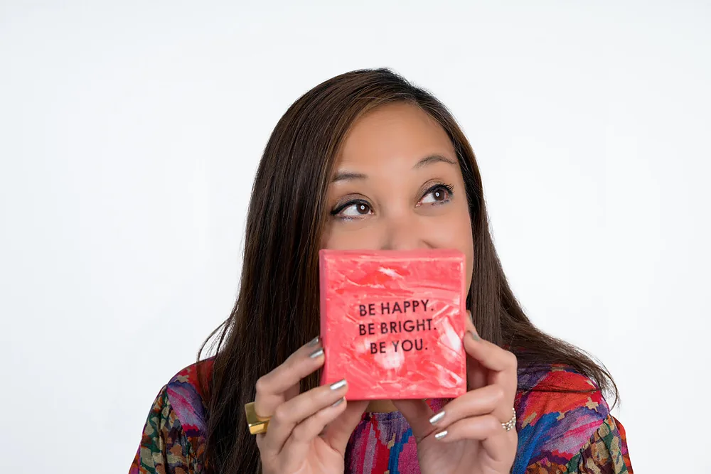 Clarissa holding one of her small paintings in front of her face. It reads: be happy, be bright.
