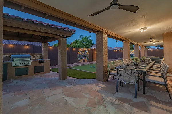A covered patio at dusk, a built in grill at one end and a long table down the middle.