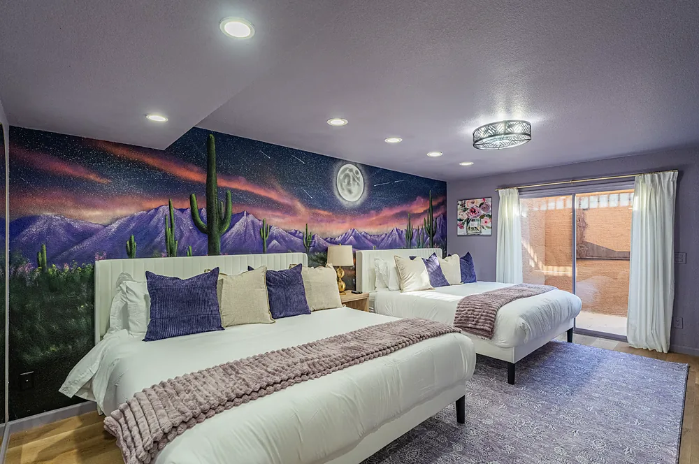 A bedroom with two beds under a hand painted mural of purple mountains, saguaros and a full moon.