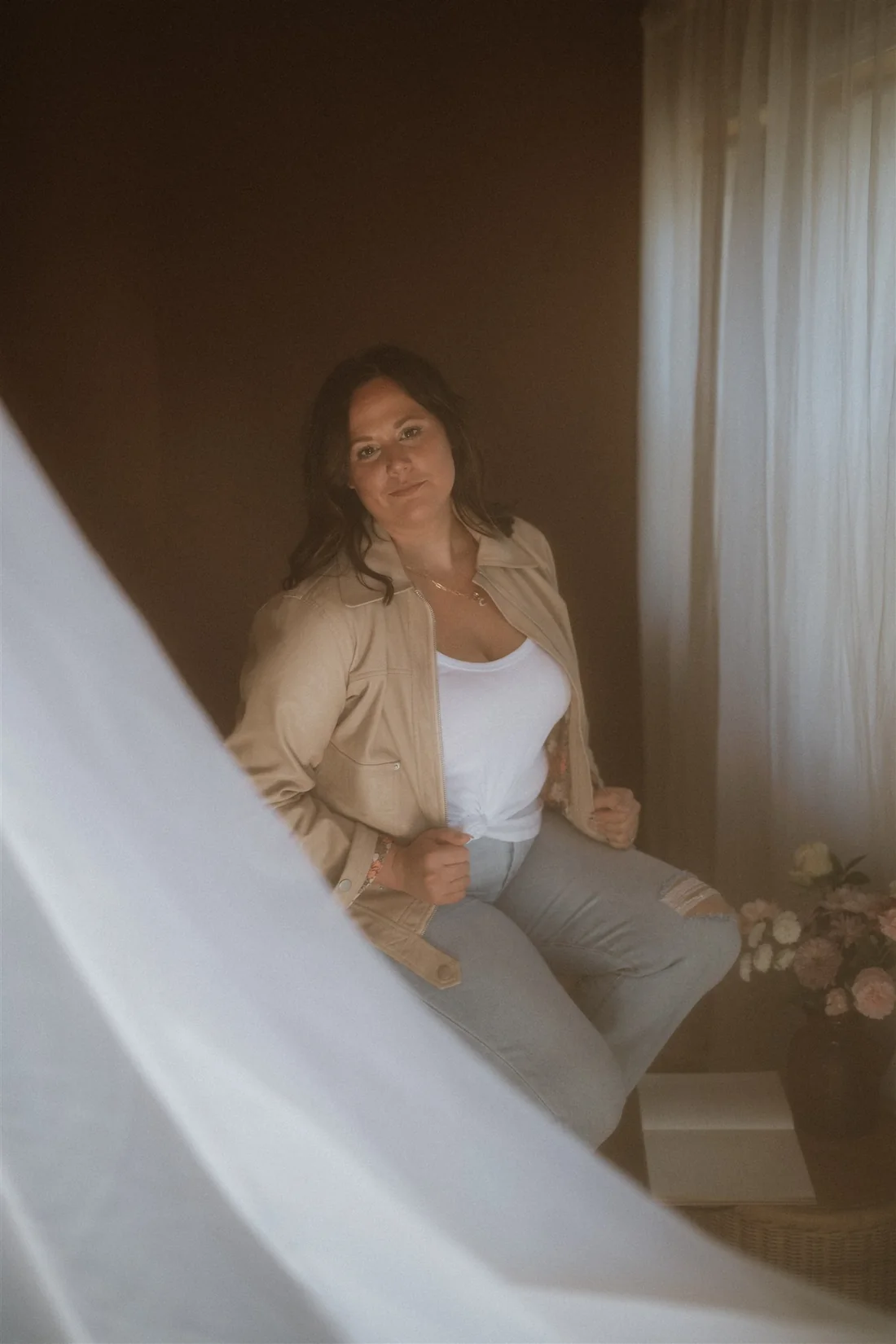 Cydnie Jocelyn seated behind a sheer curtain, half obscured by it, looking away from the camera into the light.