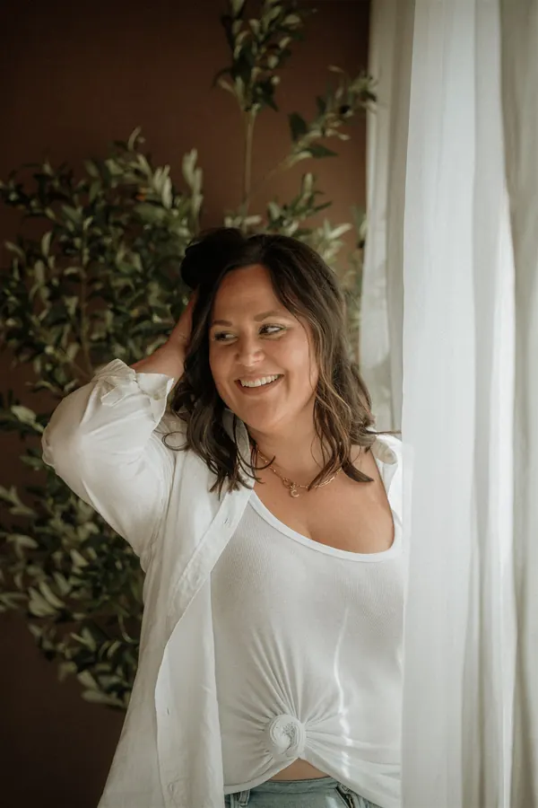 Cydnie Jocelyn, laughing by a window with her hand in her hair, in a white shirt.