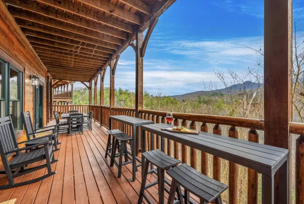 A long bar table and stools on the deck, rocking chairs beyond it and the mountains past the rail.