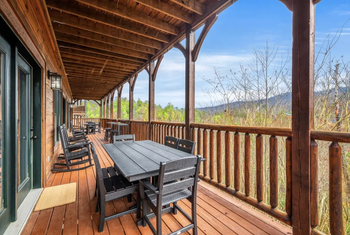A covered deck with a long table and rocking chairs, looking across bare spring woodland to a mountain ridge under a clear sky.