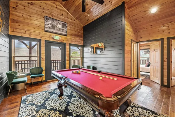 A pool table on a patterned rug, with chairs and a door onto the deck behind it.