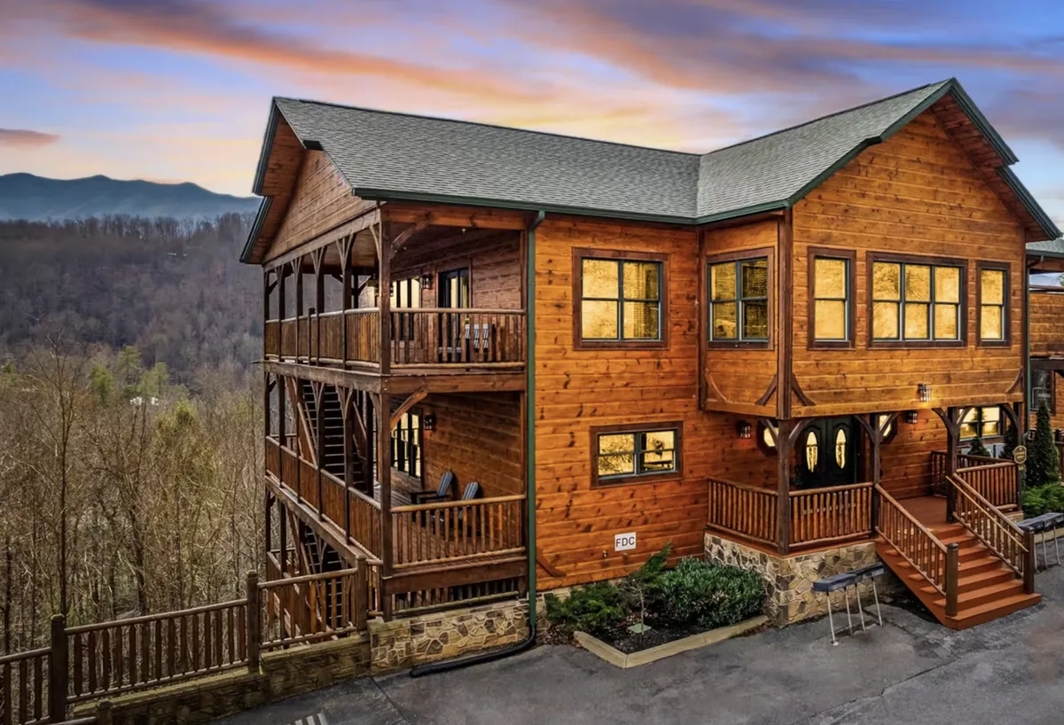 A timber house on a wooded slope at dusk, its windows lit, with the ridgeline of the Smoky Mountains behind it.