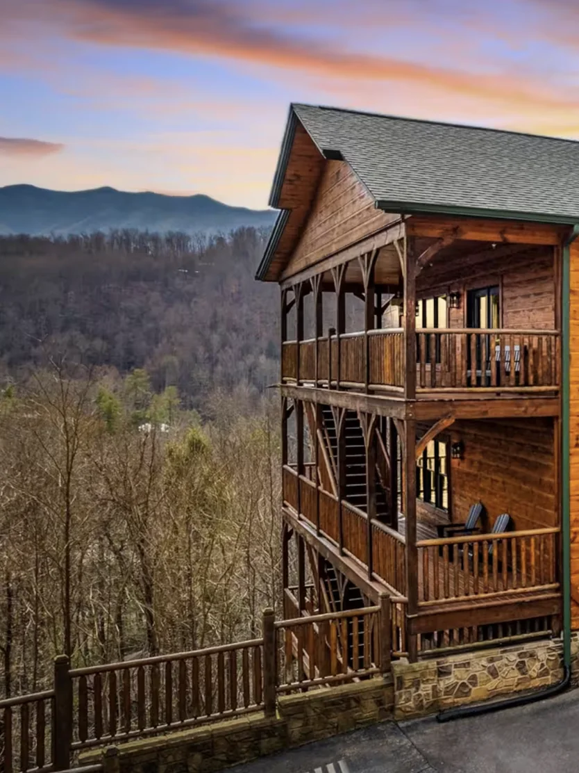 A timber house on a wooded slope at dusk, its windows lit, with the ridgeline of the Smoky Mountains behind it.