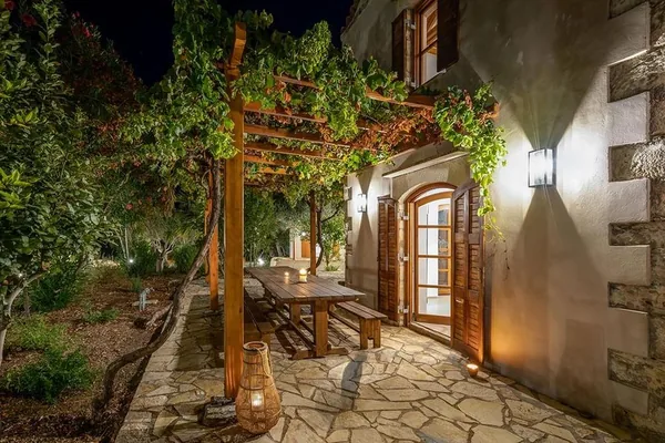The lit entrance to the house at night, a table under the vine pergola beside the door.