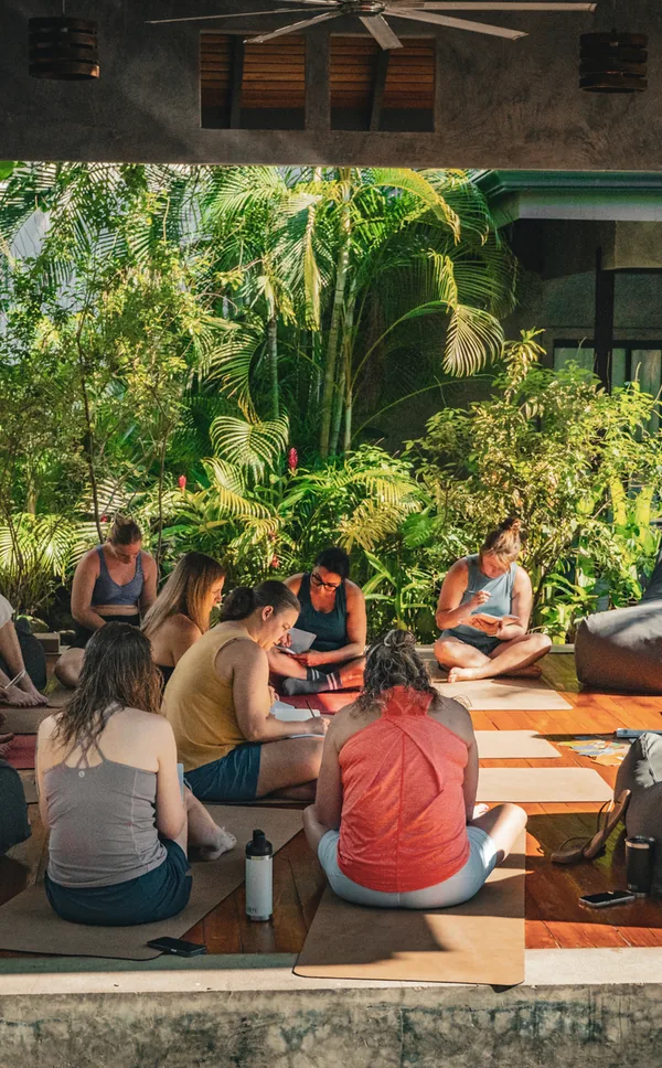 Eight women sitting on mats on an open pavilion floor, most of them seen from behind, writing in notebooks with palms and ginger flowers behind them.