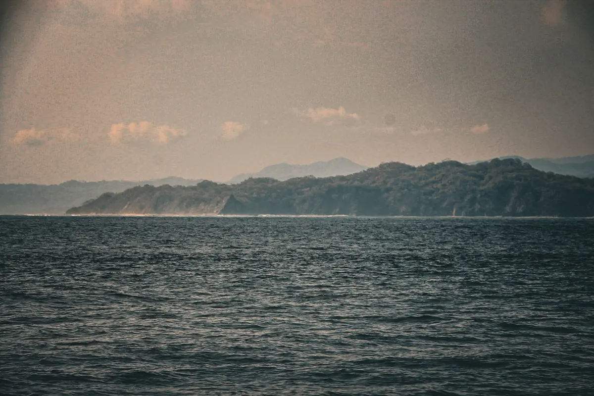 A headland across still water under a pale sky, seen from the boat off the Nicoya coast, Costa Rica.