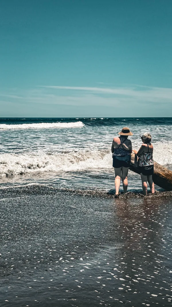 Two women standing together in the shallows with their backs to the camera, looking out at the breaking surf.